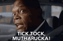 a man in a suit is driving a car and saying tick tock , mutharucka .