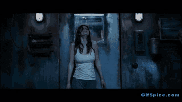 a woman in a white tank top is standing in a dark room with the website gifspice.com visible in the corner