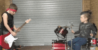 a boy with a mohawk plays drums while another boy plays a guitar