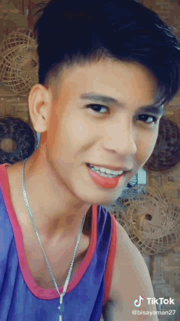 a young man wearing a purple tank top and a necklace is smiling for a tiktok video