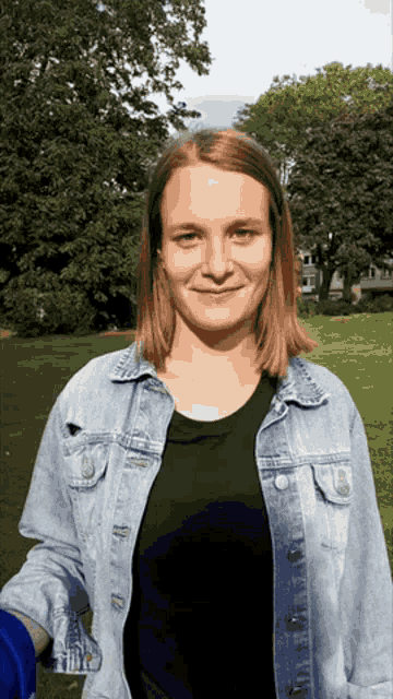 a woman wearing a denim jacket and a black shirt smiles in a park