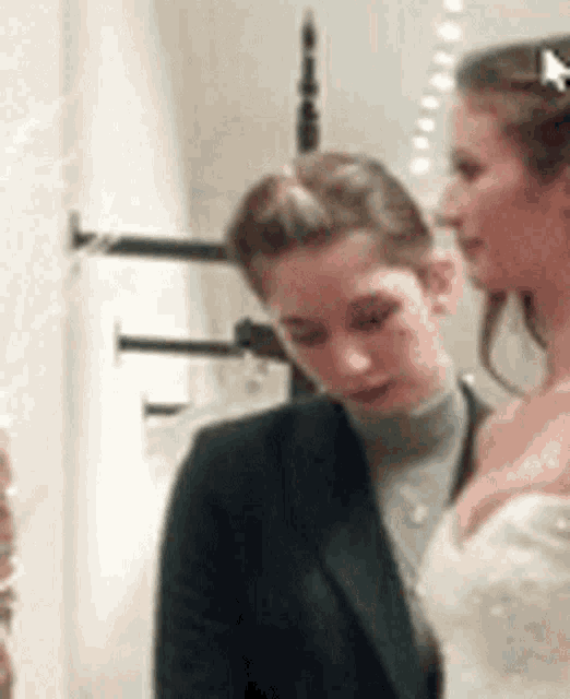 two women are standing next to each other in a room . one of the women is wearing a wedding dress .