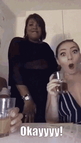 two women are sitting at a table with a glass of orange juice and the words okayyyy written on the bottom