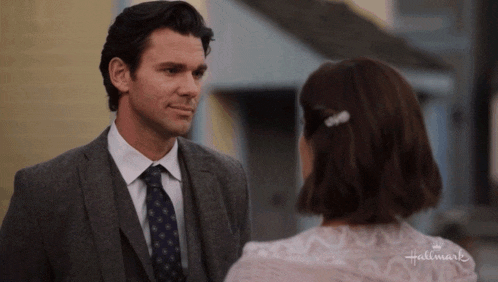 a man in a suit and tie is looking at a woman in a pink dress who is wearing a hair clip that says hallmark