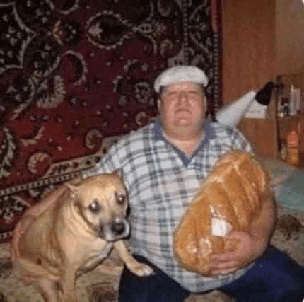 a man is sitting on a bed with a dog and holding a large loaf of bread .