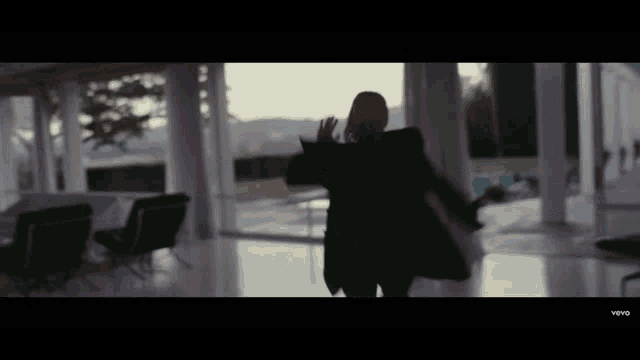 a woman in a black coat is dancing in a room with lots of windows
