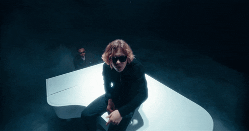 a man wearing sunglasses sits on a white grand piano