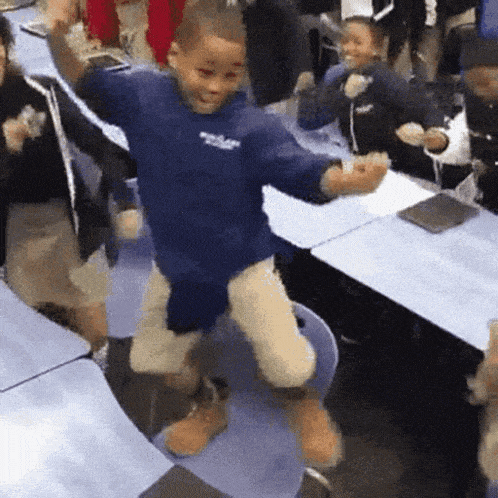 a boy in a blue shirt is jumping in the air in a classroom with other children .