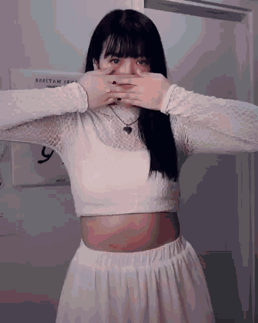 a woman wearing a white crop top and a white skirt is covering her face with her hands