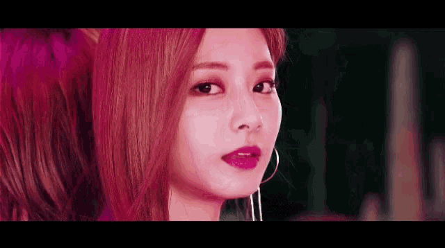 a woman with pink hair and hoop earrings looks at the camera