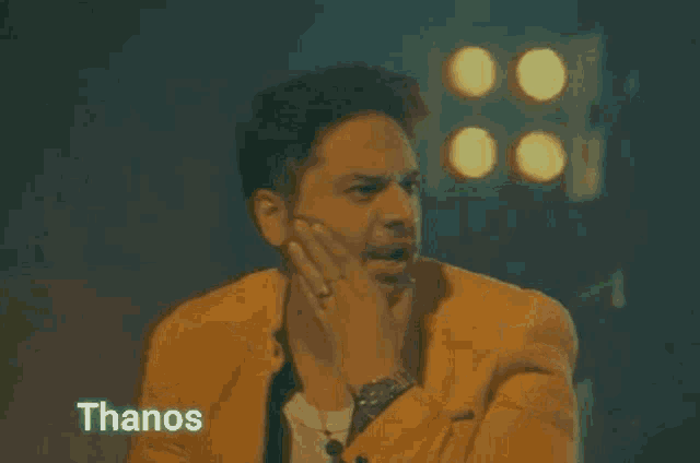 a man in a yellow suit is making a funny face and the word thanos is on the bottom right .