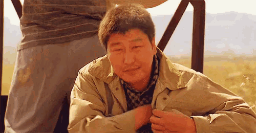 a man in a tan jacket and scarf is sitting on a bench with his hands on his chest .