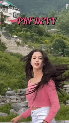 a woman in a pink crop top and white shorts is standing in front of a river with her hair blowing in the wind .