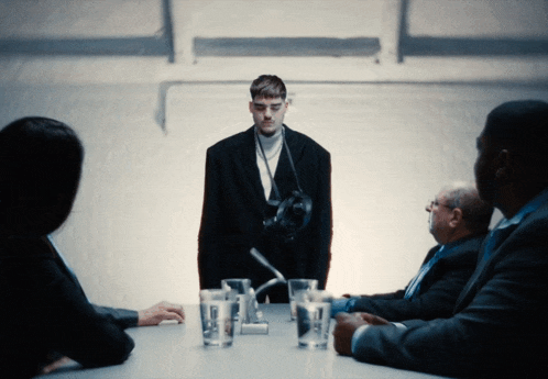 a group of people sit around a table with a man standing in front of them