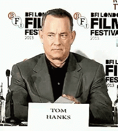 a man in a suit is sitting at a desk with a sign that says tom hanks