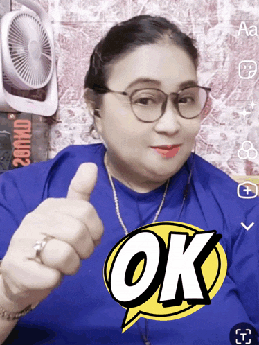 a woman wearing glasses and a blue shirt with the word ok on it gives a thumbs up