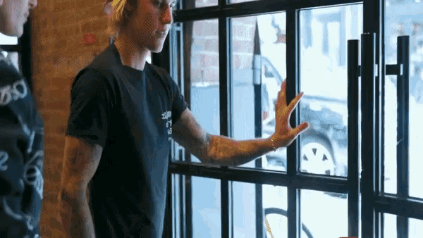 a man wearing a black shirt that says ' sd ' on it stands in front of a window