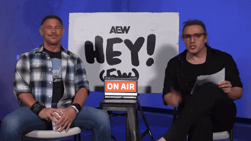 two men sit in front of a sign that says " hey "
