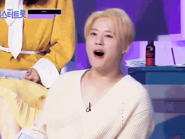 a man in a white sweater is sitting in front of a purple background with a bottle of red spray on it