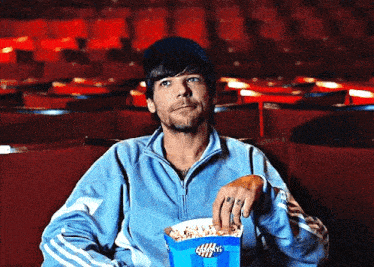 a man in a blue jacket is holding a box of popcorn in a theater .