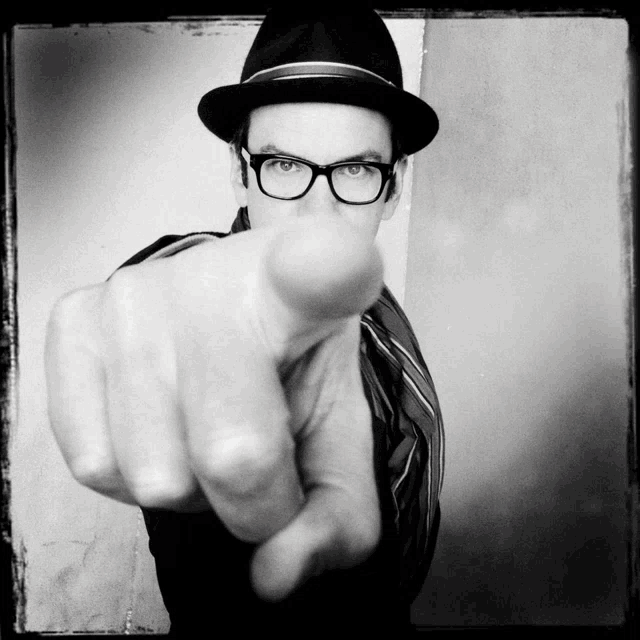 a man wearing glasses and a hat is pointing his finger at the camera