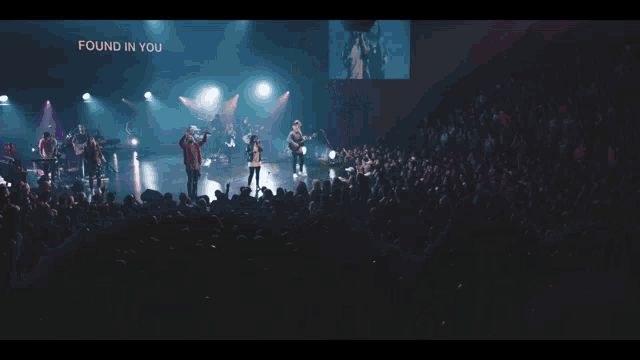 a woman singing in front of a crowd with a man playing a guitar behind her