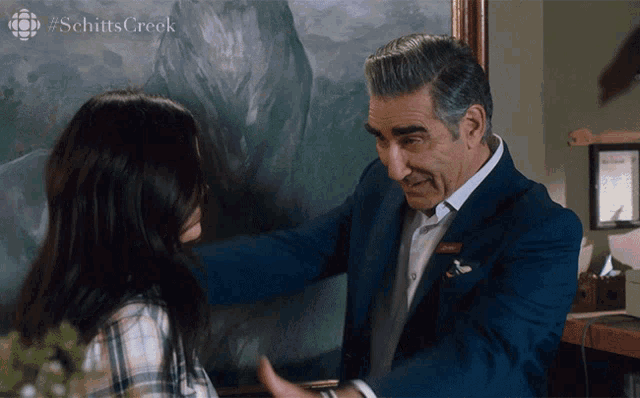 a man in a suit is hugging a woman in front of a painting with #schittscreek written on the bottom