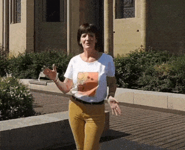 a woman in a white t-shirt and yellow pants is standing in front of a building with her arms outstretched .