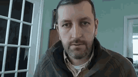 a man with a beard and a brown jacket is looking at the camera .