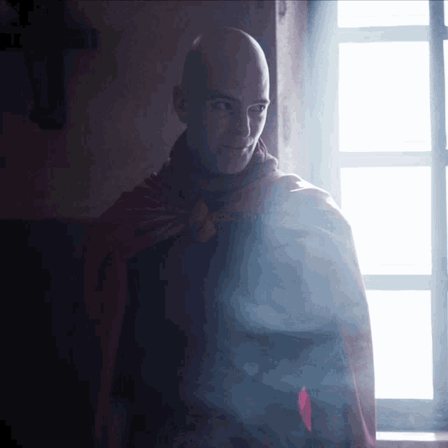 a bald man wearing a red cape is standing in front of a window