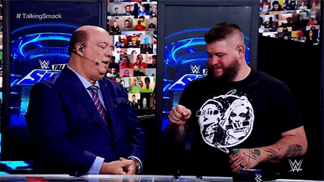 a man in a black shirt with a skull on it is talking to another man in a suit and tie .