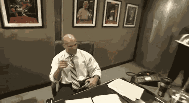 a man in a white shirt and tie sits at a desk in front of framed pictures