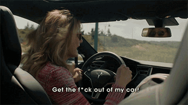 a woman is driving a tesla with the words get the f * ck out of my car