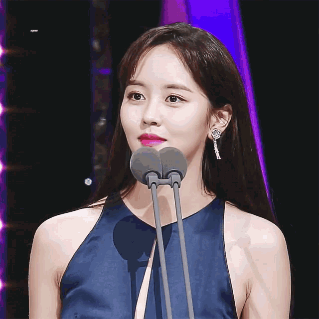 a woman in a blue dress stands in front of a microphone with a purple background behind her and the letters kpm visible