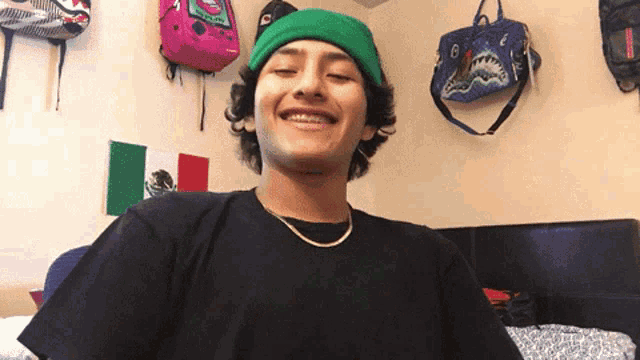 a young man wearing a green beanie and a black shirt smiles in front of a mexican flag