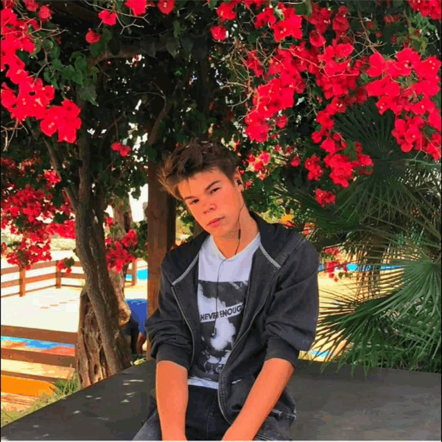 a boy wearing a never enough t-shirt sits under a tree with red flowers