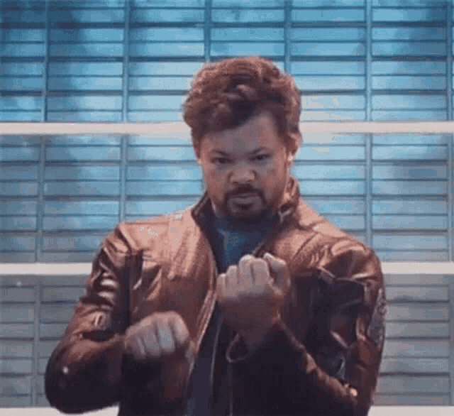 a man with a beard is wearing a leather jacket and making a fist gesture .