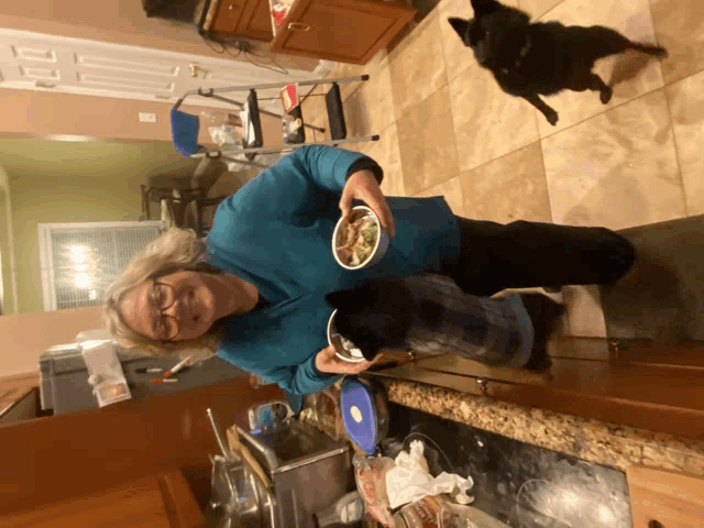 a woman in a blue shirt is holding a bowl of food while a black dog watches