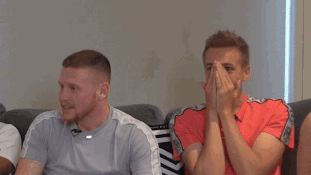 two men are sitting on a couch with their arms around each other . one of the men is wearing a red shirt .