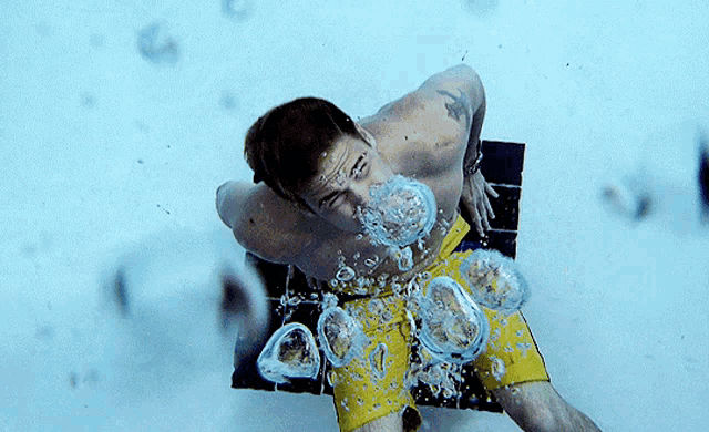 a shirtless man in yellow shorts is swimming underwater with bubbles coming out of his mouth