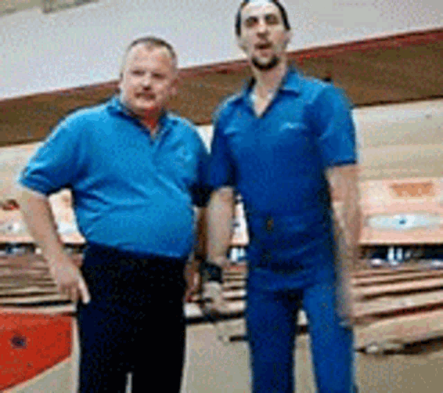 two men in blue shirts are posing for a picture in a bowling alley