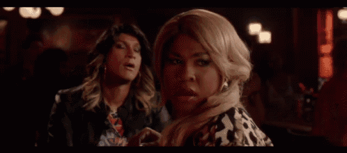 a woman in a wig is sitting next to another woman in a dark room and looking at the camera .