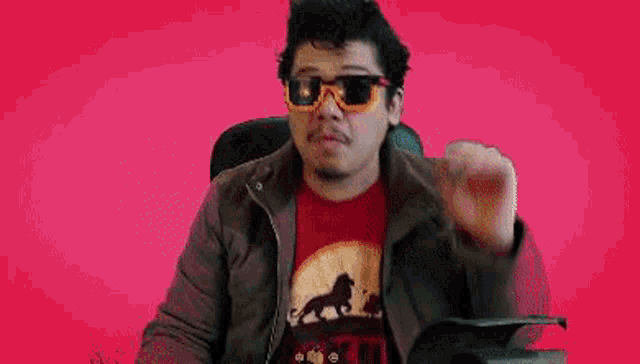 a man wearing sunglasses and a haku t-shirt is dancing on a pink background .