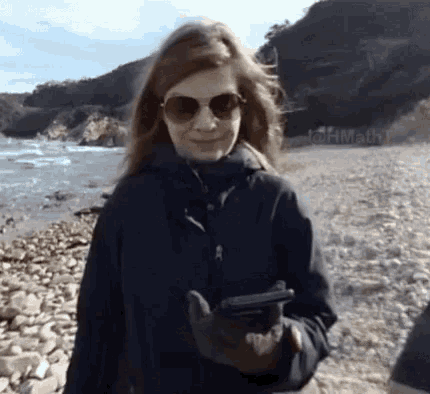 a woman wearing sunglasses and a black jacket is walking on a rocky beach holding a cell phone .