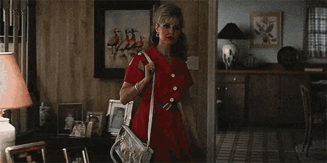 a woman in a red dress is standing in a living room holding a white purse .