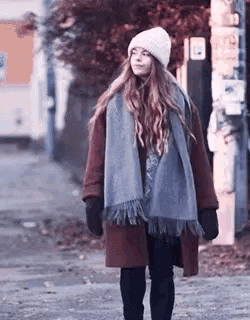 a woman wearing a scarf , coat , hat and gloves is walking down a street .