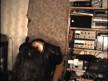 a man in a black leather jacket is standing in front of a shelf with electronics on it
