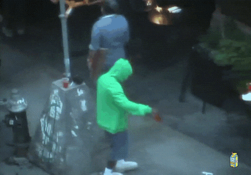 a man in a green hoodie is walking down a street with a carton of lemonade in the background