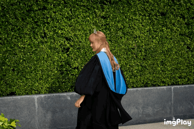 a woman in a graduation cap and gown is walking in front of a hedge with imgplay written in the corner