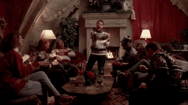 a group of people are gathered in a living room with a man in a sweater standing in the middle